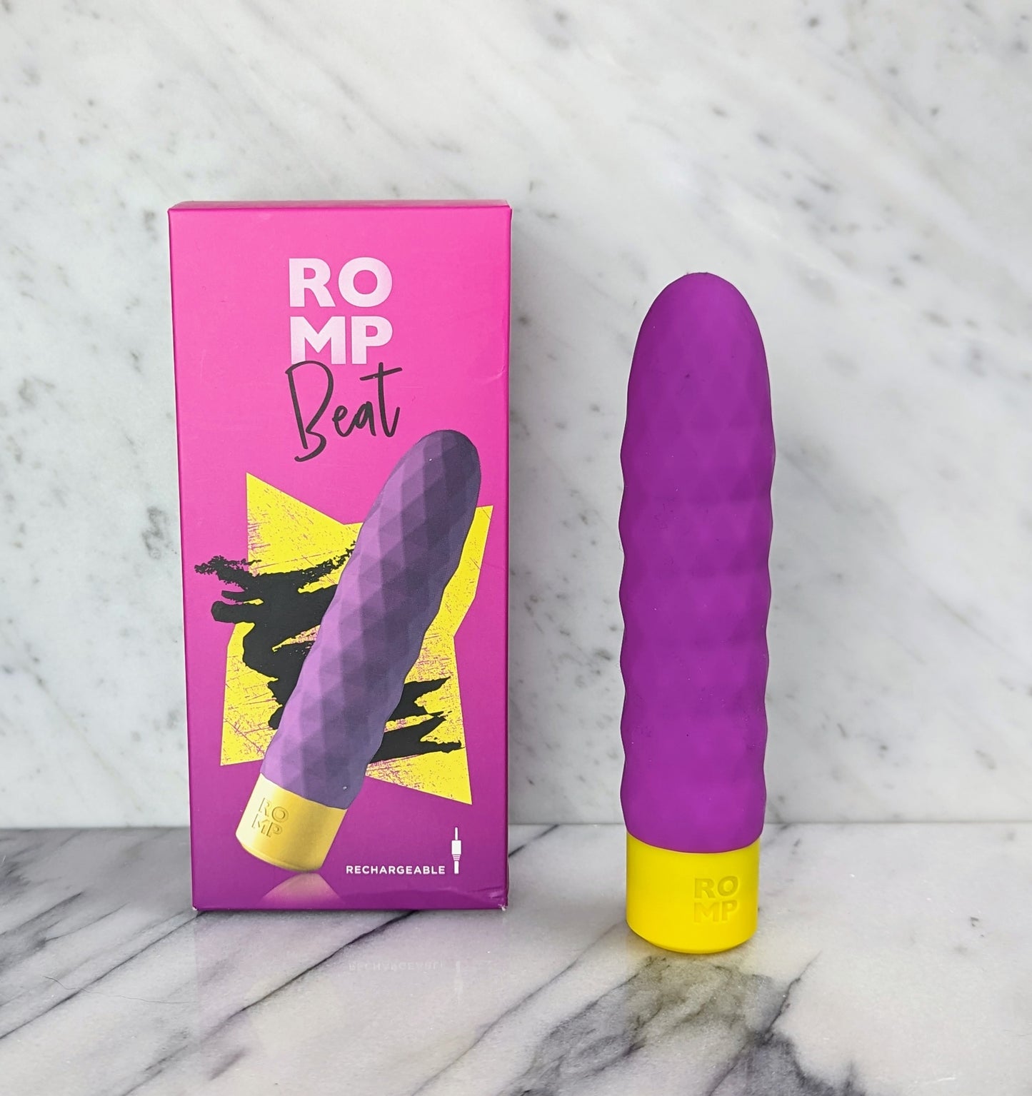 ROMP Beat vibrator in paars met gele basis naast roze productdoos op marmeren ondergrond